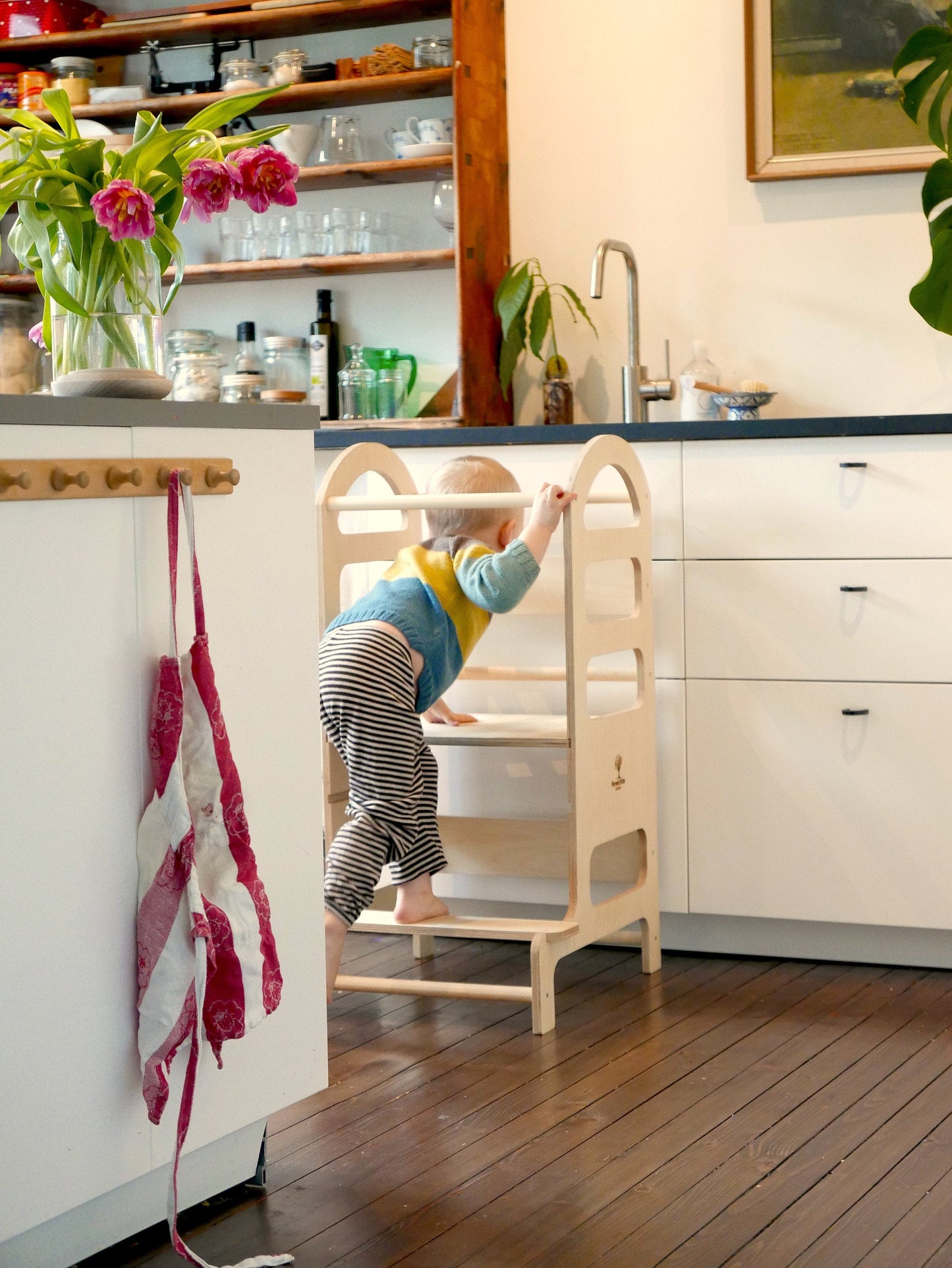 Liten gutt på cirka 1,5 år som klatrer opp på et Montessori læringstårn i tre på et lyst kjøkken – norsk kjøkkenhjelper for barn til trygg lek og læring