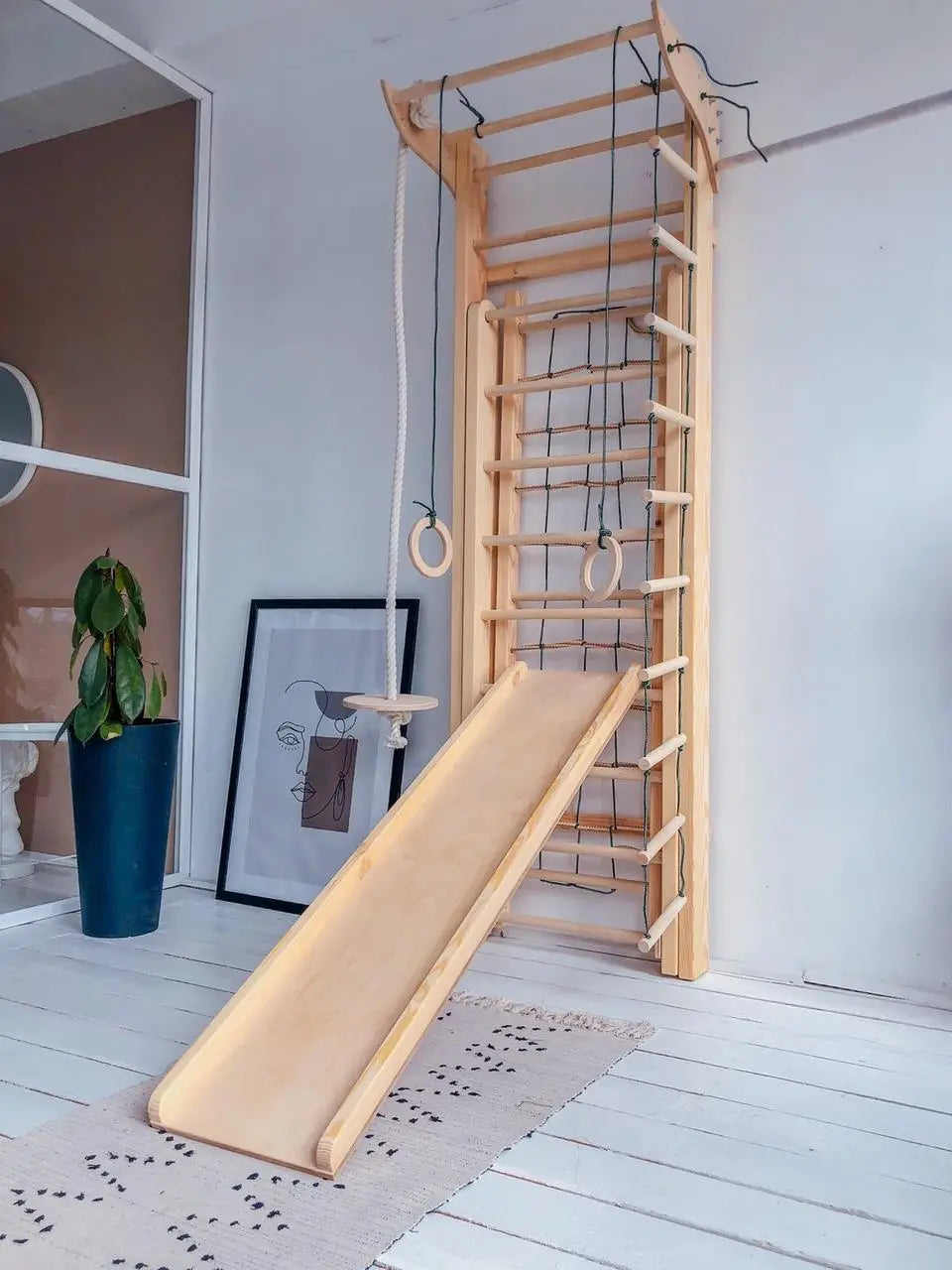 A wooden indoor gymnastics and play structure with a ladder, hanging rings, a small platform, and a slide, shown in a bright room with white walls and floorboards.