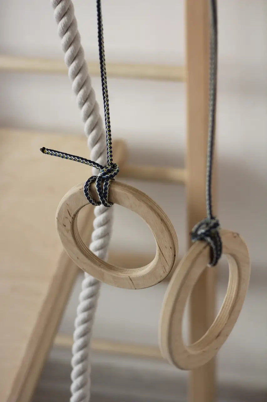 Two natural wood gymnastic rings hanging from a wooden frame, with one ring attached to a white rope and the other to a dark braided rope, shown in a softly lit indoor setting.