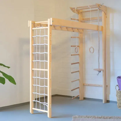 A light natural wood children's gymnastics and climbing wall with multiple accessories, including a rope ladder, wooden rings, a hanging platform, and a rope swing, set against a white paneled wall.