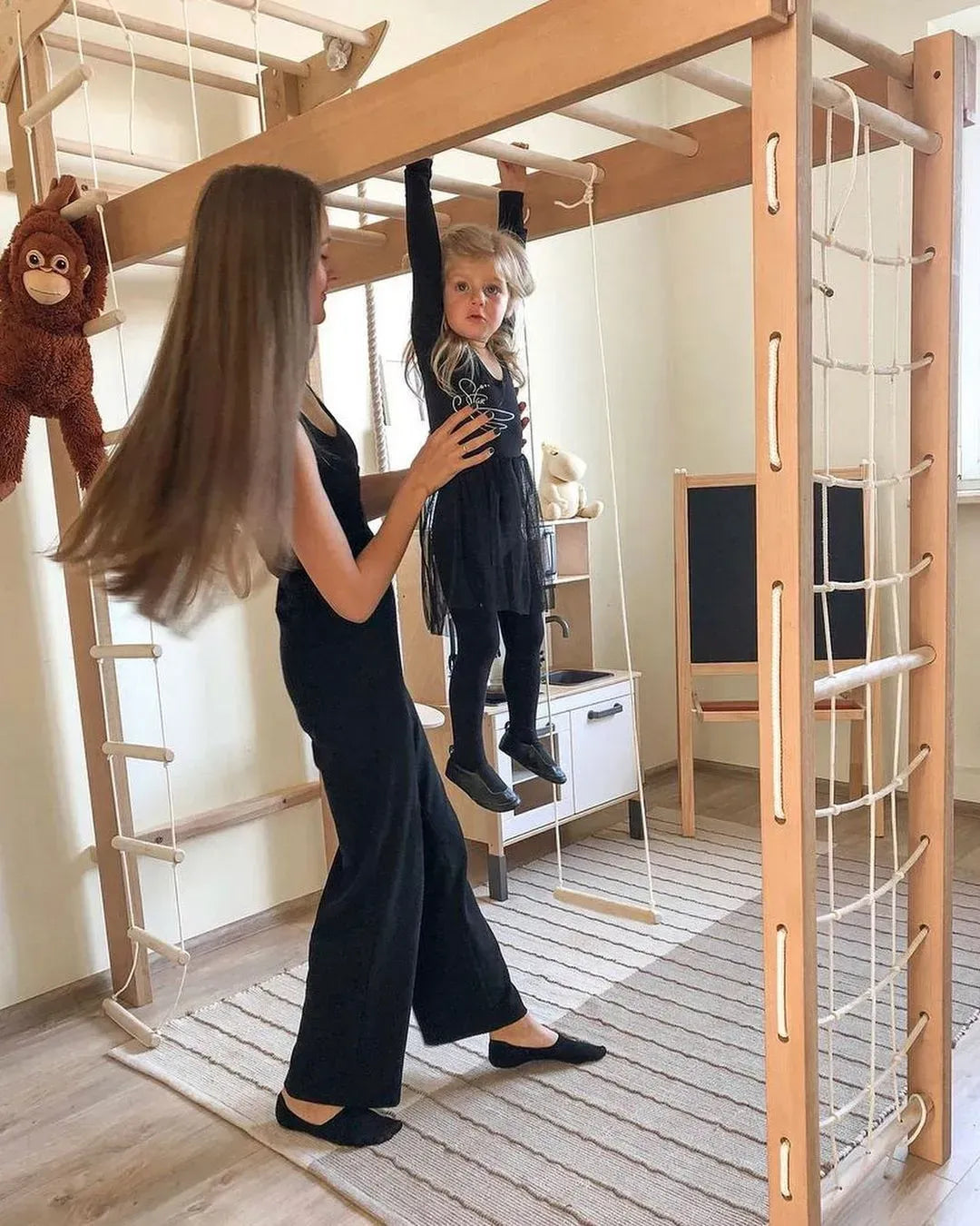 A woman in a black dress assists a young child wearing a black outfit while the child hangs on a wooden climbing frame with ropes and bars in a bright indoor playroom.