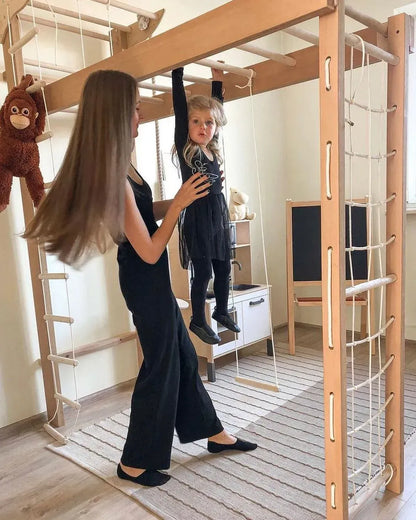 A woman in a black dress assists a young child wearing a black outfit while the child hangs on a wooden climbing frame with ropes and bars in a bright indoor playroom.