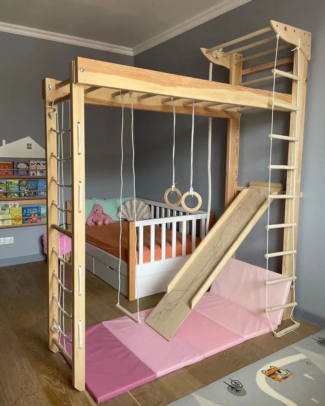 A large wooden children's play gym and loft bed frame in a bedroom, featuring a wooden slide, climbing ladder, rope rings, hanging ropes, and a pink foam mat on the floor.