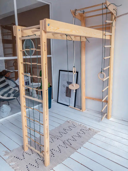 A large natural wood children's climbing gym with multiple climbing walls, rings, and a hanging disc, set up on a white wooden floor with a patterned rug.