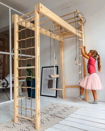 A young girl in a pink t-shirt and light pink skirt plays on a large wooden multi-functional gym structure with a ladder, rope climbing wall, and hanging rings, set in a bright room with white wooden floors.