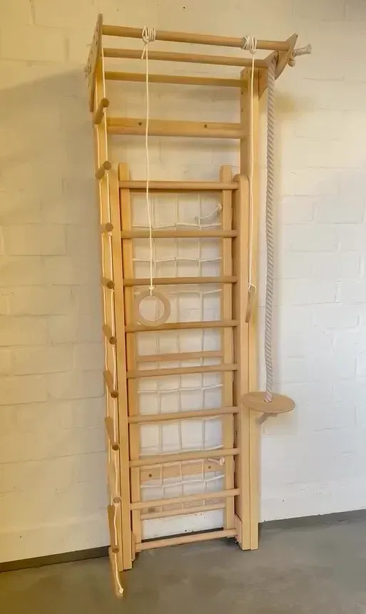 A tall wooden climbing wall with multiple rungs, a hanging rope, a small circular ring, and a side platform, photographed against a white brick wall.