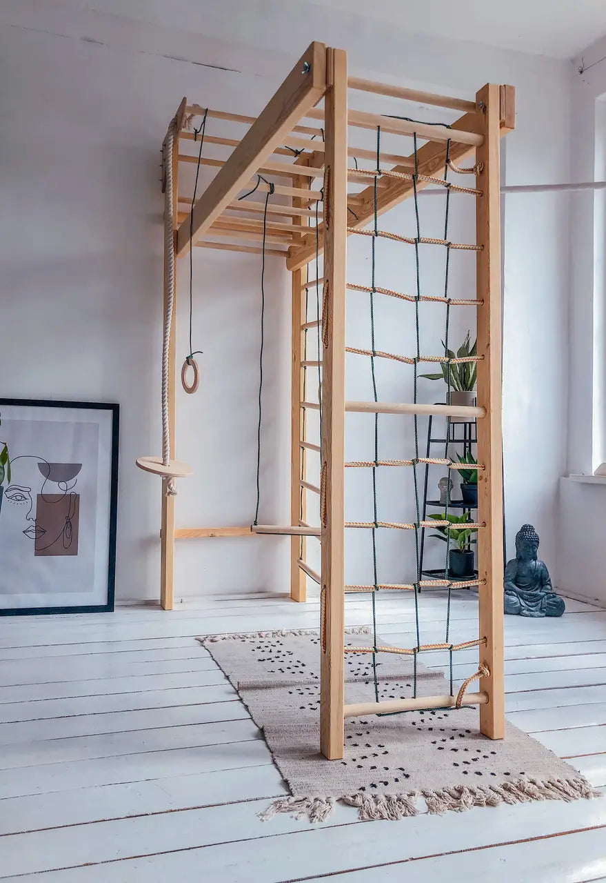 A wooden multi-functional climbing gym structure with a ladder, rings, rope, and catwalk, set up on a white wood floor with a beige rug in a bright room.