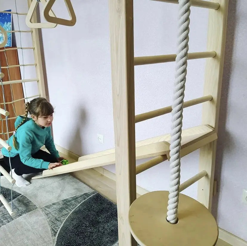 A young girl in a teal sweatshirt and black pants is kneeling on a gray patterned floor next to a large wooden children's climbing wall with a thick rope. The room has light lavender walls and a white electrical outlet.