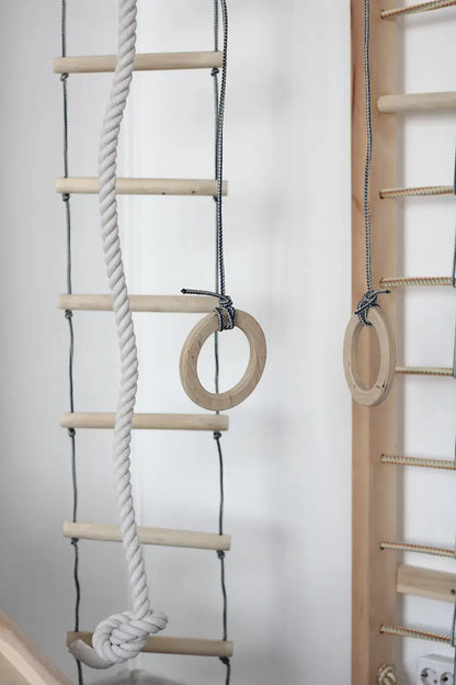 Wooden wall climbing ladder with natural wood rungs, a thick white rope, and hanging wooden rings against a white wall.