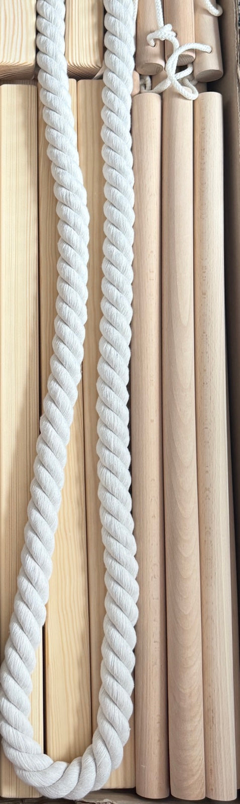 A close-up of a thick, white, twisted rope in front of a stack of light natural wood dowels or poles.