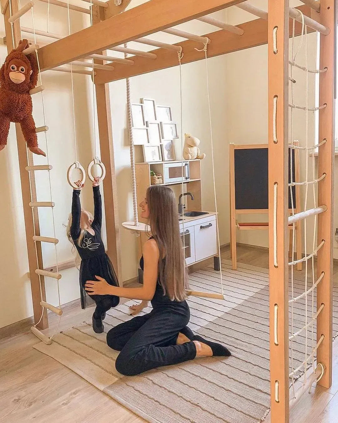 A woman kneels on a striped rug in a wooden playroom, supporting a young child hanging from gymnastic rings. The room features a large wooden climbing frame with ropes, a monkey hanging from the left, a small white play kitchen, and a chalkboard.