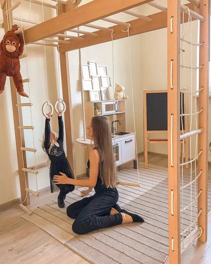 A woman kneels on a striped rug in a wooden playroom, supporting a young child hanging from gymnastic rings. The room features a large wooden climbing frame with ropes, a monkey hanging from the left, a small white play kitchen, and a chalkboard.